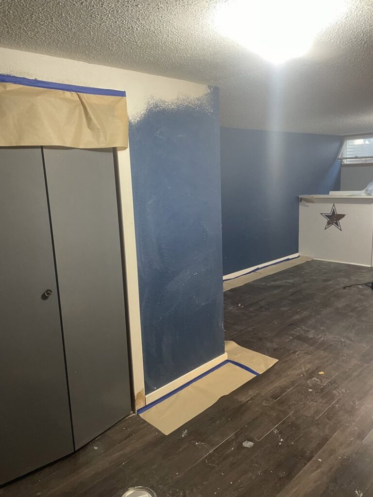 An interior wall being painted dark blue, with masking tape and paper protecting the floor, by Knutson's Property Maintenance Llc in Williston, ND.