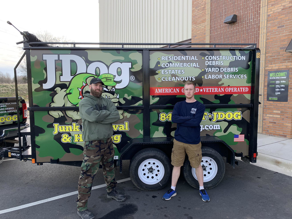 Two team members from JDog Junk Removal & Hauling Concord, NC, standing proudly in front of their branded junk removal trailer.