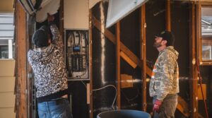 Two Junk Commandos team members demolishing a wall, exposing electrical components, during a junk removal project in Tacoma, WA.