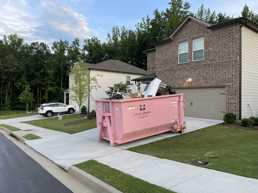 A pink dumpster from Little Junkers filled with various junk items in a residential driveway in Peachtree City, GA.