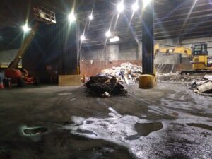 Inside a large junk processing facility with piles of waste, a yellow excavator, and a scissor lift, managed by Lemcor in Newark, NJ.