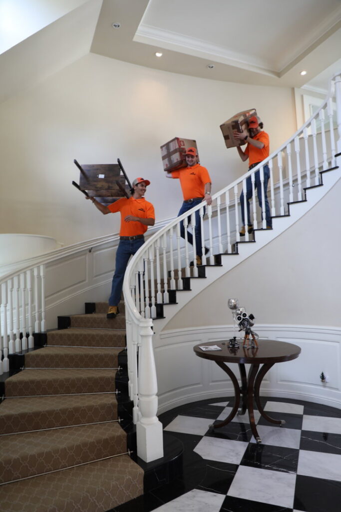 The Junkiez Junk Removal crew carrying boxes and a chair down a grand staircase inside a home in Los Angeles, CA.