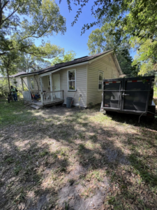 A junk removal dump trailer parked beside a house with a worker, provided by Just Dump It in Jacksonville, FL.
