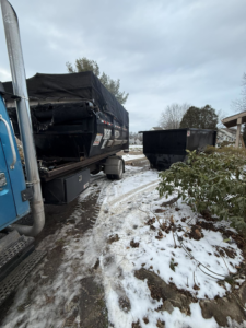 A blue truck delivering a black dumpster for junk removal services in a snowy residential area by Wagners Property Services LLC in Canton, OH.