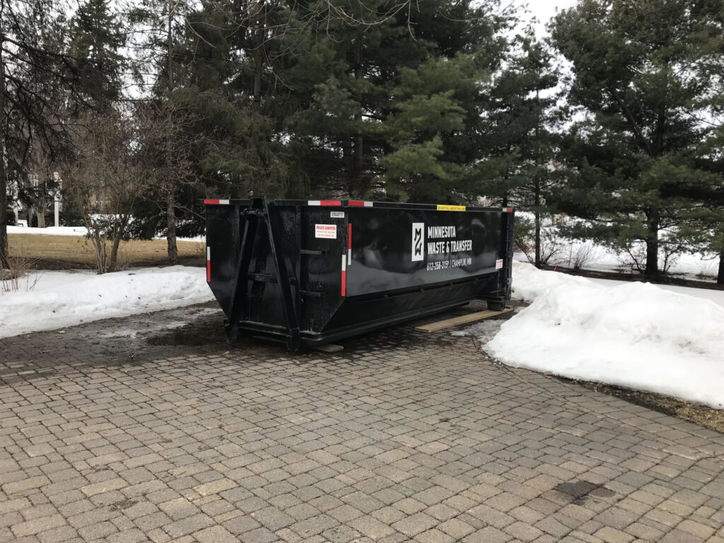 A large black junk removal dumpster from Minnesota Waste and Transfer placed on a paved driveway in Champlin, MN during winter