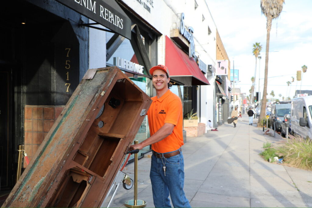 A The Junkiez Junk Removal specialist moving a large wooden cabinet on a hand truck down a city sidewalk in Los Angeles, CA.
