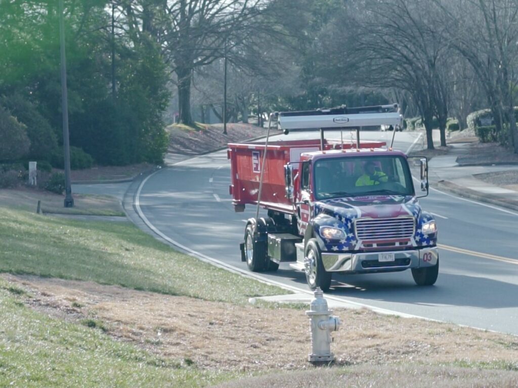 A Waste Removal USA truck with a red roll-off dumpster driving on a road for general junk removal services in Austin, TX