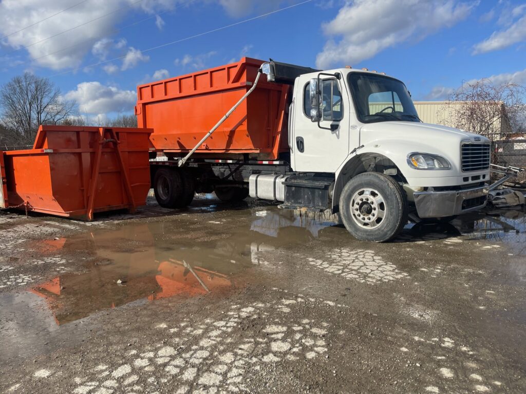 A junk removal truck with an orange dumpster and another empty dumpster in a muddy lot by Lil Man W/A Can LLC in Columbus, OH.