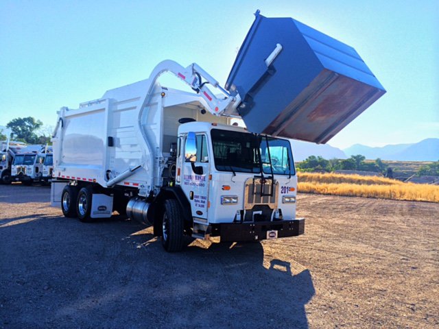 A white junk removal truck from ECONO WASTE INC in Ogden, UT, lifting a large blue dumpster with its hydraulic arm.