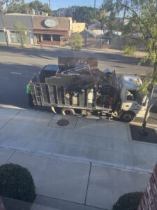 A junk removal truck parked on a street, loaded with items, with a worker at the back, by Larry The Rubbish Man in Los Angeles, CA.