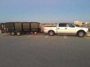 A silver pickup truck with a trailer full of black junk removal bins at dusk for Industry Services, LLC in Inglewood, CA.