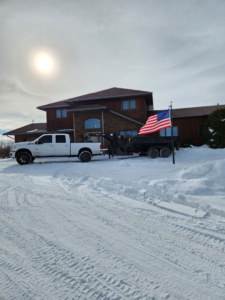 A white Ford truck with a dump trailer parked near a house with an American flag, ready for junk removal by Outlaw Rental in Bismarck, ND.