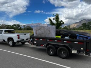 A Junk Solutions pickup truck towing a trailer loaded with a mattress and other junk on a road in Ogden, UT, showcasing a completed removal job.