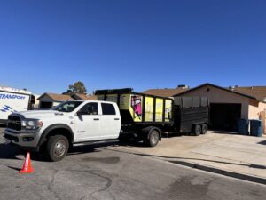 A junk removal truck and trailer with a branded dumpster at a residential job site for Las Vegas Hauling Junk & Moving in Las Vegas, NV