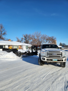 A white Ford truck with a dump trailer parked in front of a snowy house, ready for junk removal by Outlaw Rental in Bismarck, ND.