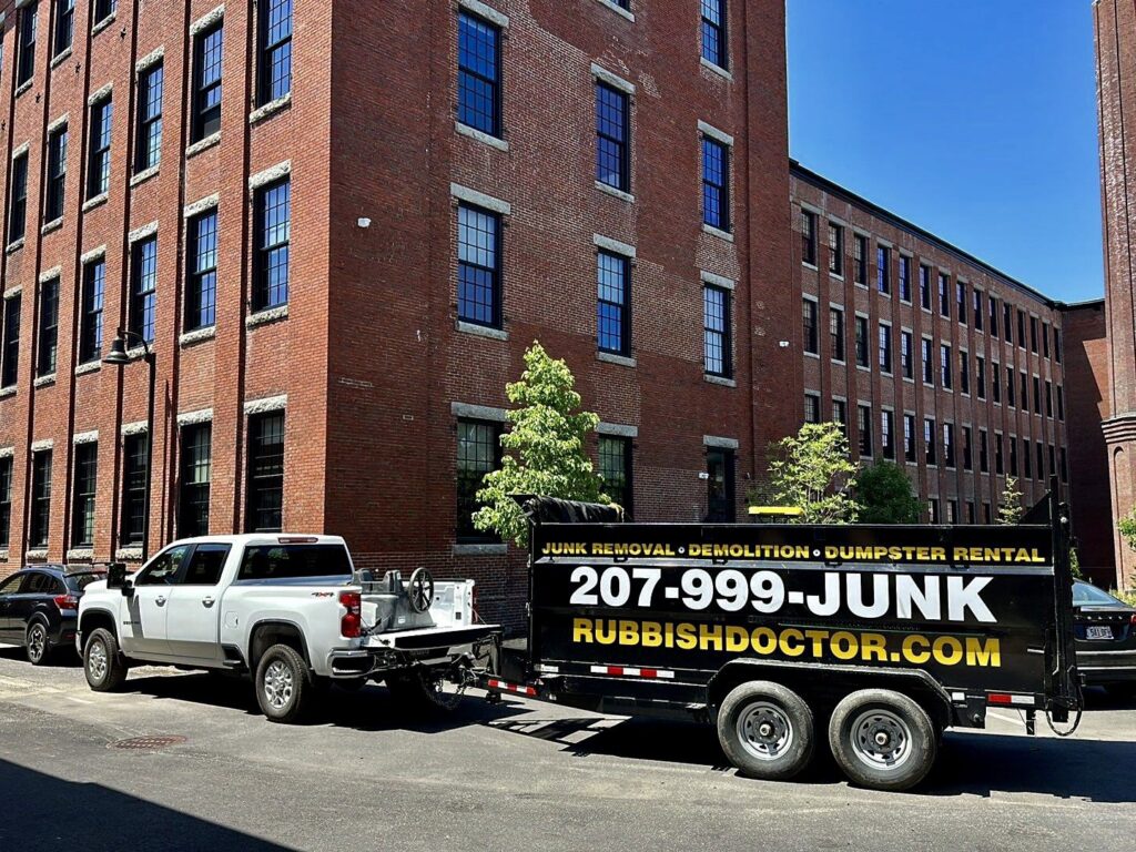 A Rubbish Doctor branded truck and trailer parked on an urban street in front of brick buildings in Portland, ME.
