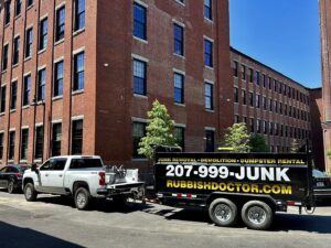 A Rubbish Doctor branded truck and trailer parked on an urban street in front of brick buildings in Portland, ME.