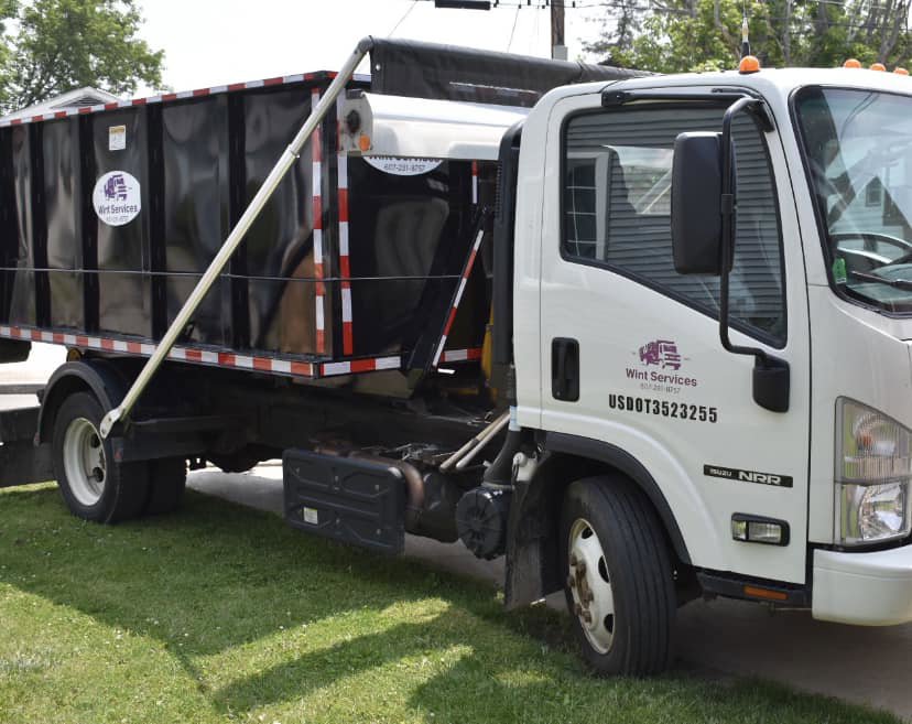 A white junk removal truck with a black roll-off dumpster from Wint Services, LLC, parked on a lawn in Colorado Springs, CO.