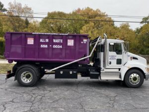 An All In Waste junk removal truck with a purple dumpster loaded on the back, ready for service in Greenville, SC.