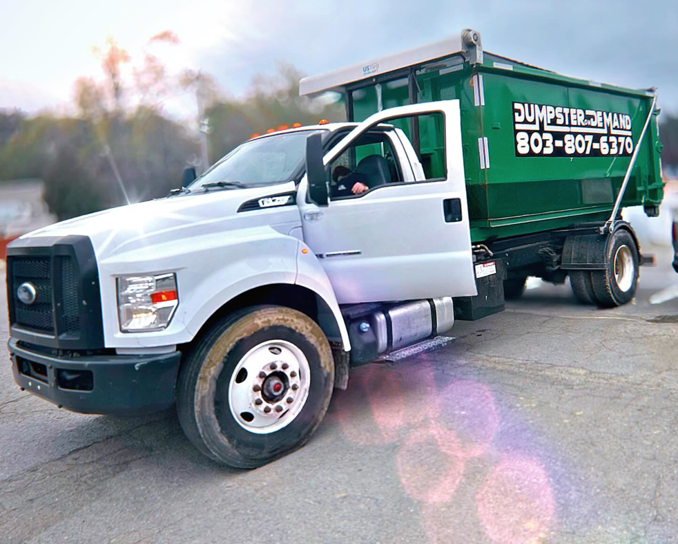 A white junk removal truck carrying a green dumpster from Dumpster on Demand in Columbia, SC.