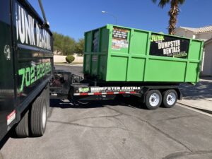 A Junk Control junk removal truck pulling a trailer with a green dumpster on a residential street in Henderson, NV.