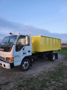 A white Isuzu junk removal truck with a yellow dumpster from Dump N Dash Dumpsters LLC, parked on a rural road in Valley, NE.