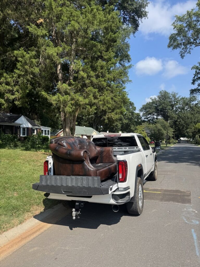 A Junkjet pickup truck hauling a large brown armchair on a residential street in Charlotte, NC.