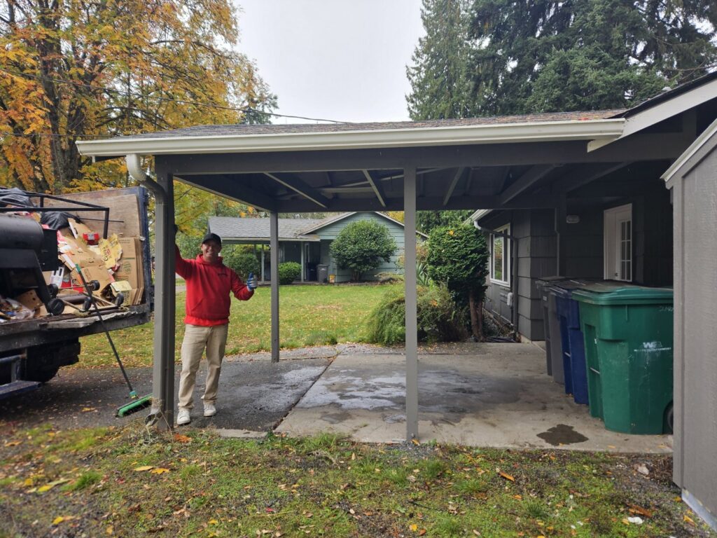 A Junkzilla Inc. worker giving a thumbs-up next to a partially loaded junk removal truck at a residential carport in Everett, WA.