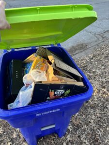A blue KC Disposal residential trash bin filled with cardboard and household waste in Kansas City, KS.