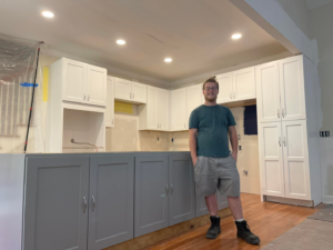 A handyman stands proudly in a kitchen with newly installed white and grey cabinets during a renovation by Wanderworks in Charleston, SC.
