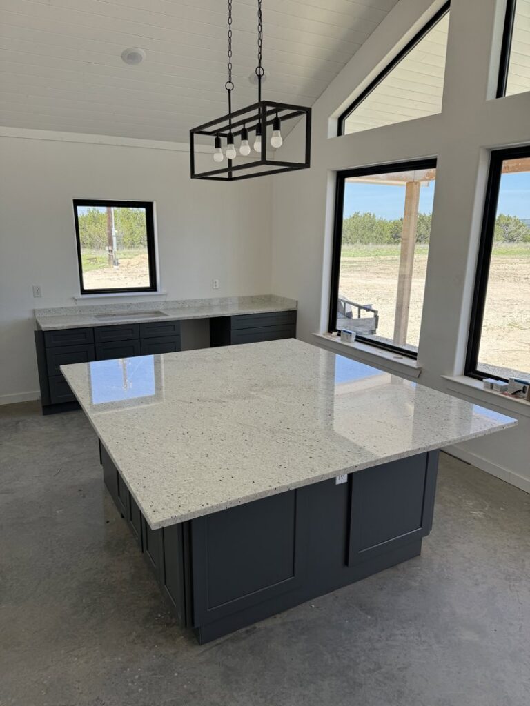 A kitchen construction in progress with dark gray cabinets and a large white island by Stone Age Granite of Austin, TX.