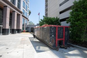 A large black roll-off dumpster placed next to a modern commercial building by Waste Master Dumpster Rental Orlando, FL.