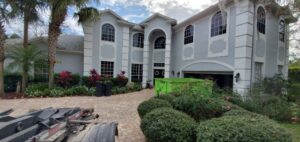A green dumpster being delivered to a large residential driveway for junk removal by Bin There Dump That Dumpster Rental Orlando, FL.