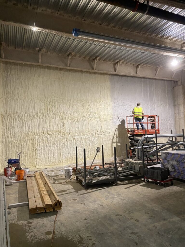 A worker on a scissor lift applying spray foam insulation to a large interior wall in a commercial construction project by Olympus Spray Foam in Philadelphia, PA.