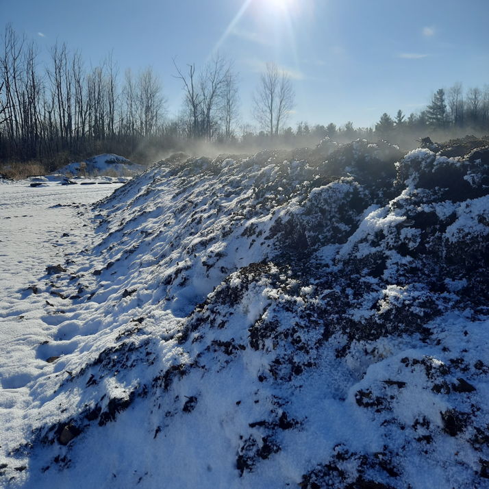 A large outdoor organic compost pile steaming in the snow, managed by Big Lake Organics in Ashland, WI.