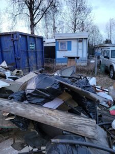 A large pile of black trash bags, wood, and various debris next to a blue dumpster, for junk removal by Alaska Waste in Anchorage, AK.