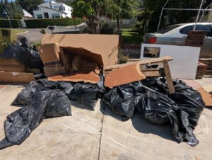 A large pile of black trash bags, cardboard boxes, and other debris on a residential driveway, awaiting removal by Larry The Rubbish Man in Los Angeles, CA.