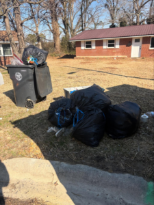 Several large black trash bags and a full bin of waste piled in a yard for pickup by JD Junk Removal in Raleigh, NC.