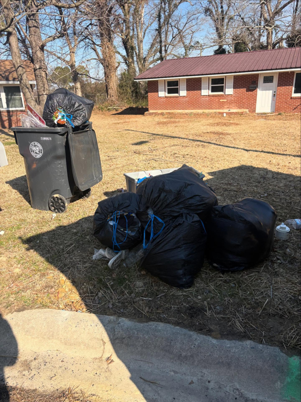 Several large black trash bags and a full bin of waste piled in a yard for pickup by JD Junk Removal in Raleigh, NC.