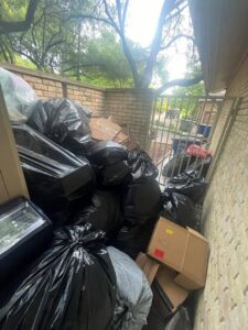 A large pile of black trash bags and cardboard boxes in an outdoor alleyway, ready for removal by Cowtown Waste Removal in Fort Worth, TX.