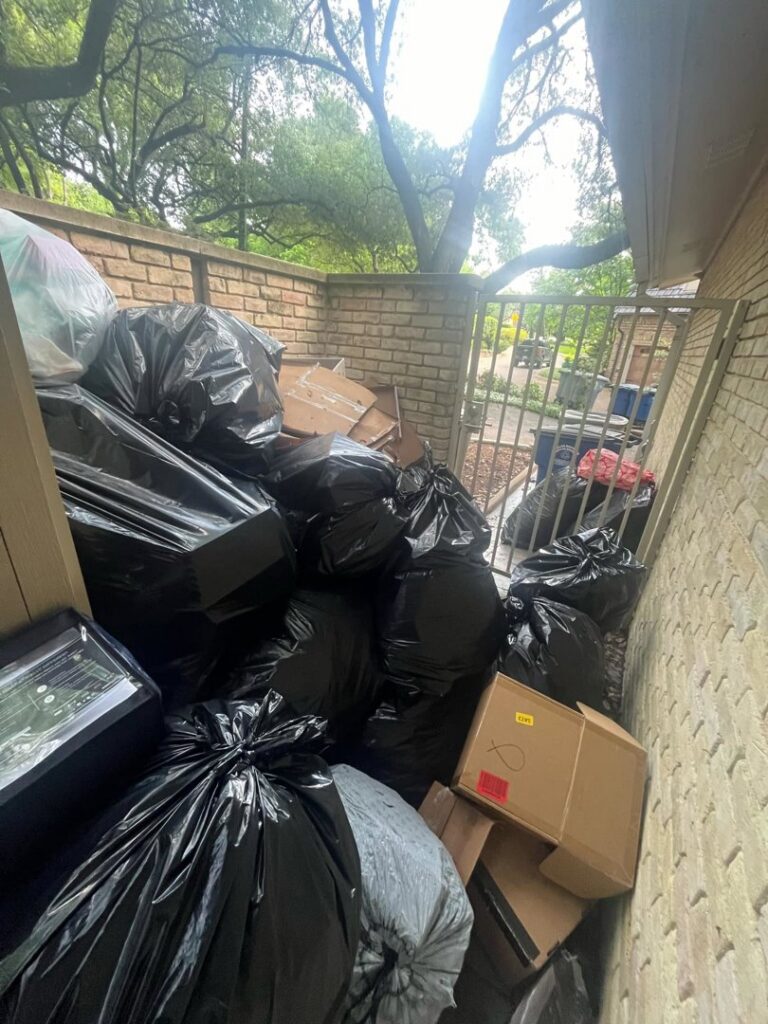 A large pile of black trash bags and cardboard boxes in an outdoor alleyway, ready for removal by Cowtown Waste Removal in Fort Worth, TX.