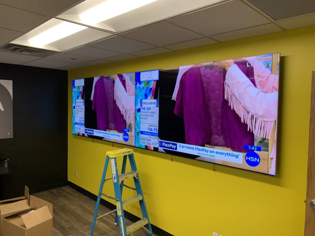 A large television being mounted on a yellow wall, a commercial service by I Know A Guy Handyman Service, Cottage Grove, MN.