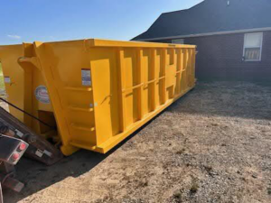 A large yellow roll-off dumpster placed on a gravel area next to a residential building by Tecumseh Rolloff Services in Moore, OK.