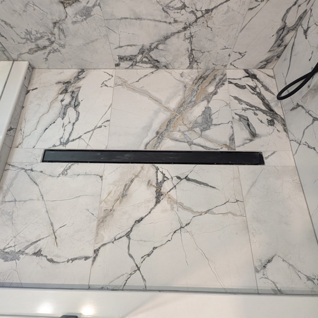 A sleek linear shower drain installed in a marble-look tiled shower floor by Ensotile - Atlanta Bathroom Remodeling in Stone Mountain, GA