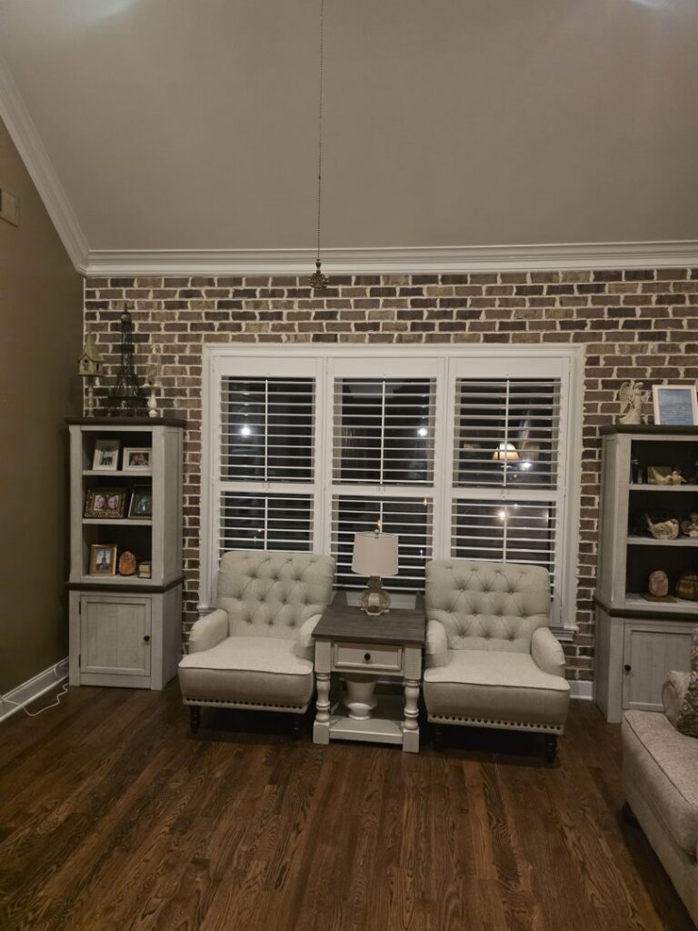 A living room featuring a brick accent wall and white plantation shutters installed by CMT Construction LLC in Columbia, MO.
