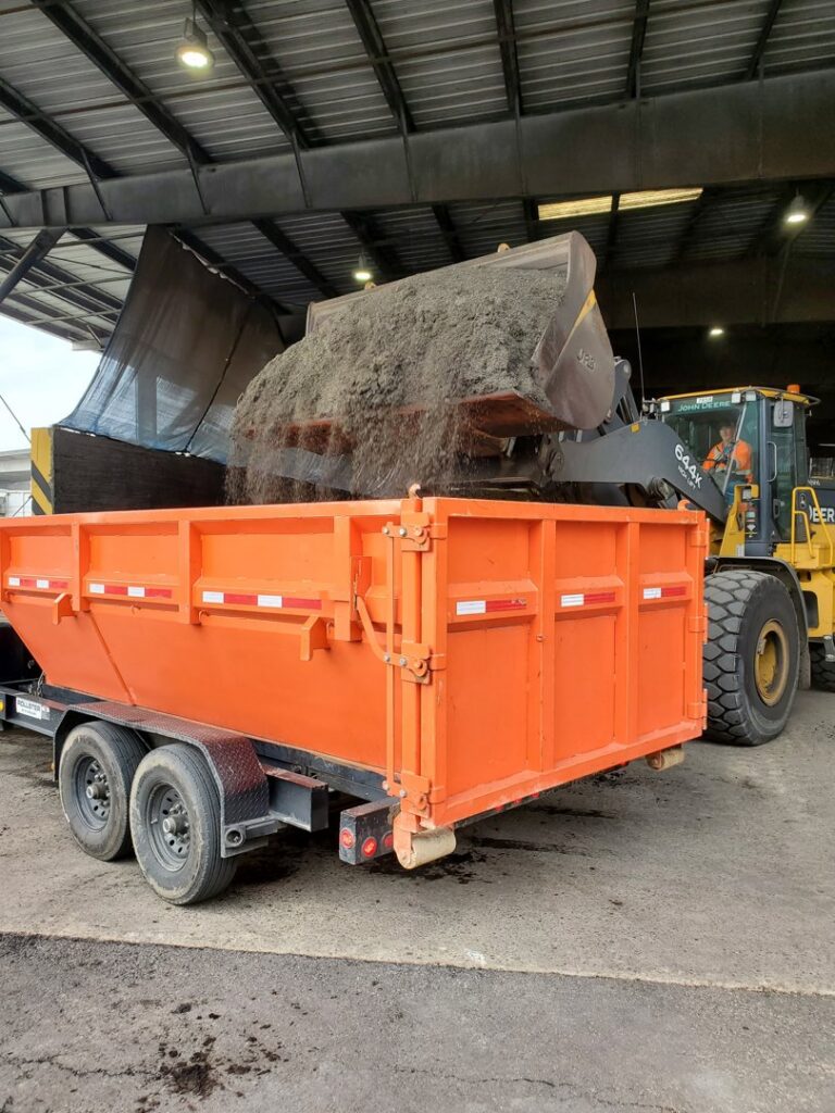 A front-end loader dumping debris into an orange roll-off dumpster at a transfer station for ADE Hauling in Tacoma, WA