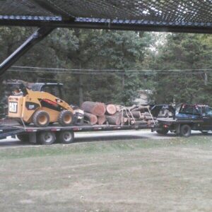 A trailer loaded with logs and a skid steer being transported, showcasing logistics for KT Farms, Tree Service & Logging LLC in Springfield, MO.
