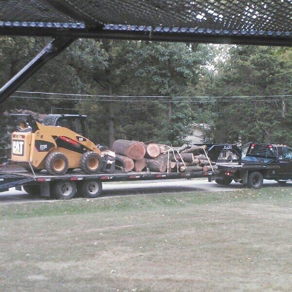 A trailer loaded with logs and a skid steer being transported, showcasing logistics for KT Farms, Tree Service & Logging LLC in Springfield, MO.