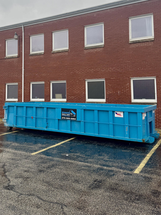 A long blue roll-off dumpster from Ryan's Roll-Off Dumpsters parked in a wet parking lot in Owensboro, KY.