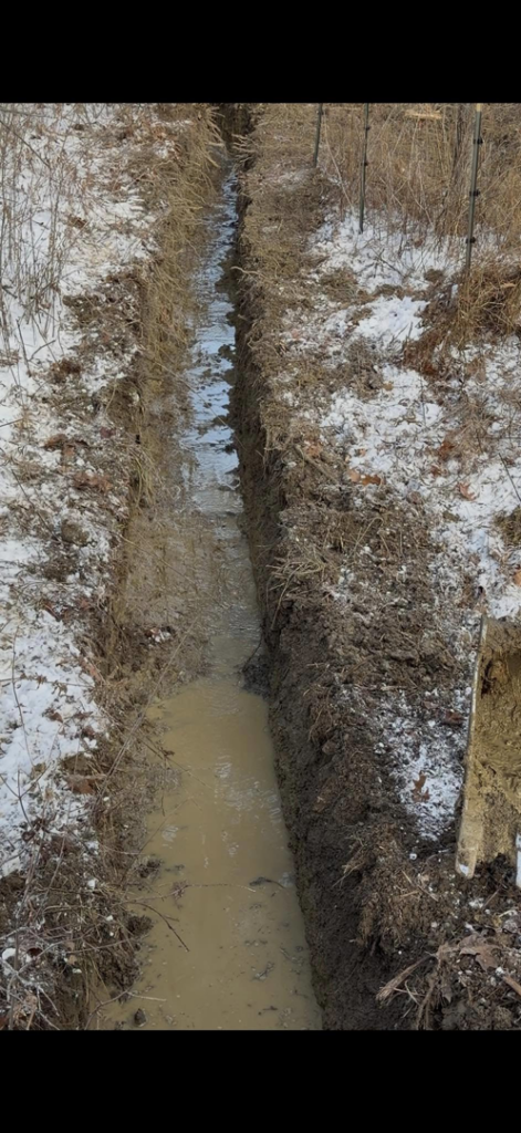 A long excavated trench filled with muddy water, indicating drainage work by Naz General Contracting LLC in Schenectady, NY.